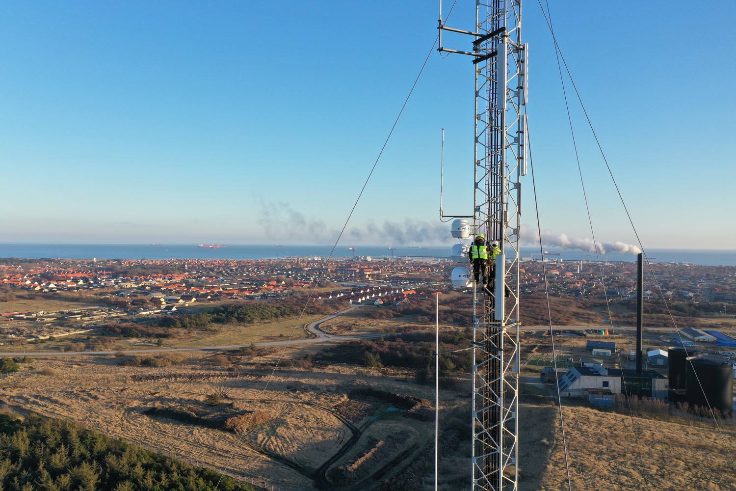 A technician climbing a Nowhere Networks shore-to-ship radio mast above a coastal town, with the sea on the horizon.